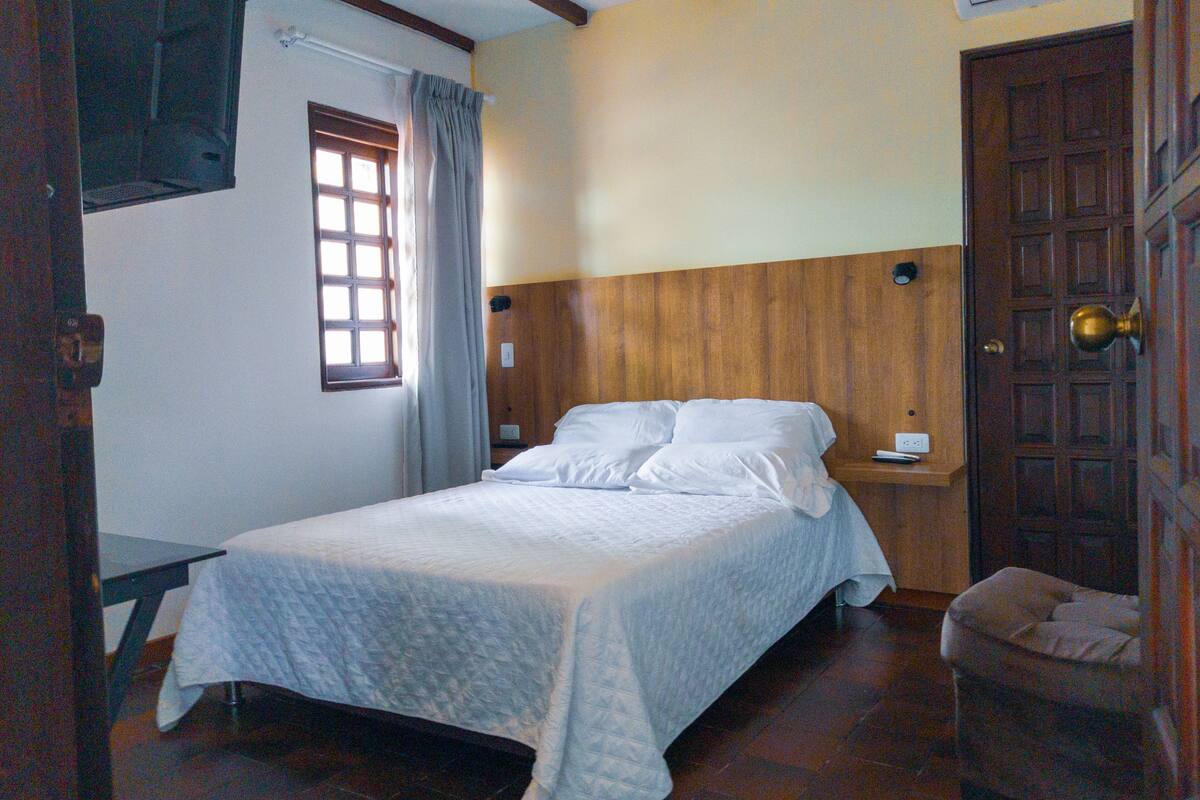 Un letto rifatto con cura in una stanza con travi a vista, una finestra con tende, una porta in legno e un comodino.