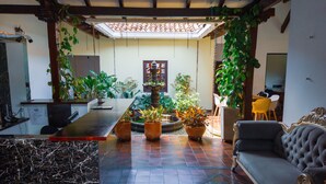 Een ruime kamer met een lange eettafel, diverse potplanten, een comfortabele bank en een kleine fontein.