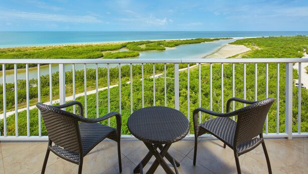 Un balcon avec deux fauteuils en osier et une petite table, donnant sur un paysage côtier verdoyant, avec une rivière.
