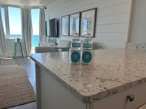 A kitchen with a marble countertop, two water bottles, a teal chair, and a view of the ocean through large windows.