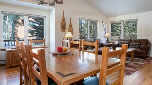 Amplia sala de estar con una mesa de comedor de madera, sillas y un sofá. Grandes ventanales ofrecen vistas al exterior.