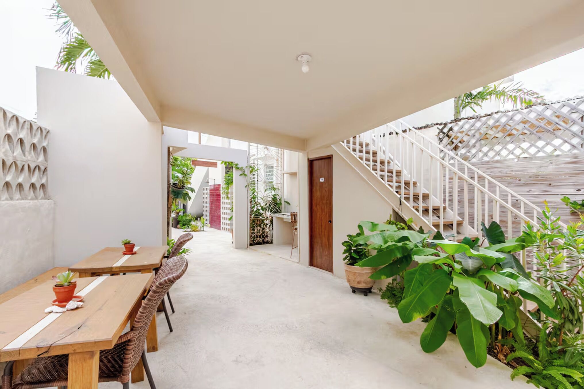 Un espacio al aire libre techado con una mesa y sillas de madera, plantas en macetas y una escalera blanca.