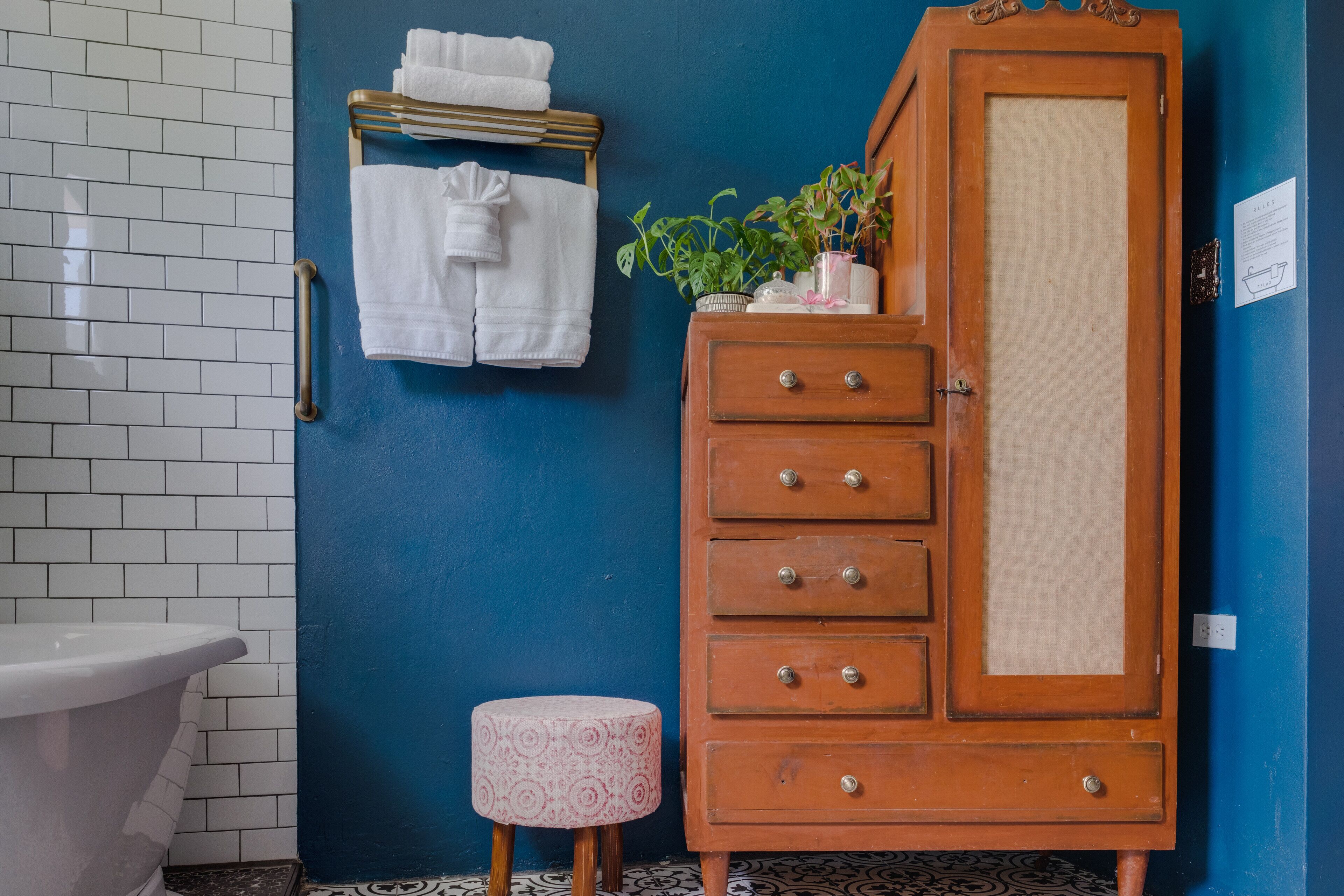 A bathroom with a white toilet, a blue wall, a wooden cabinet with drawers, a small stool, and a towel rack with towels.