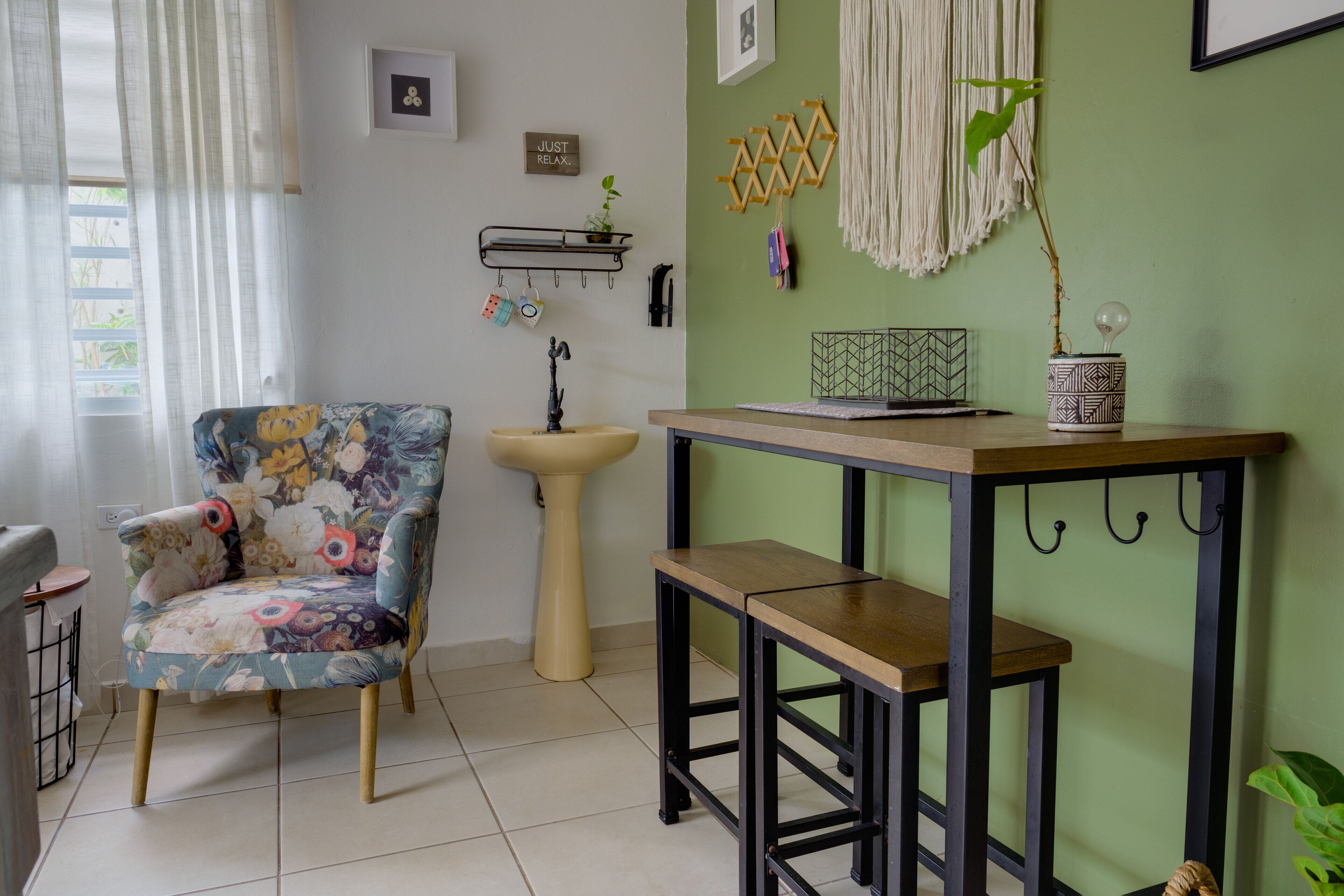 Una habitación con un sillón floral, una mesa de madera con un banco, un lavabo y una estantería de pared con objetos decorativos.