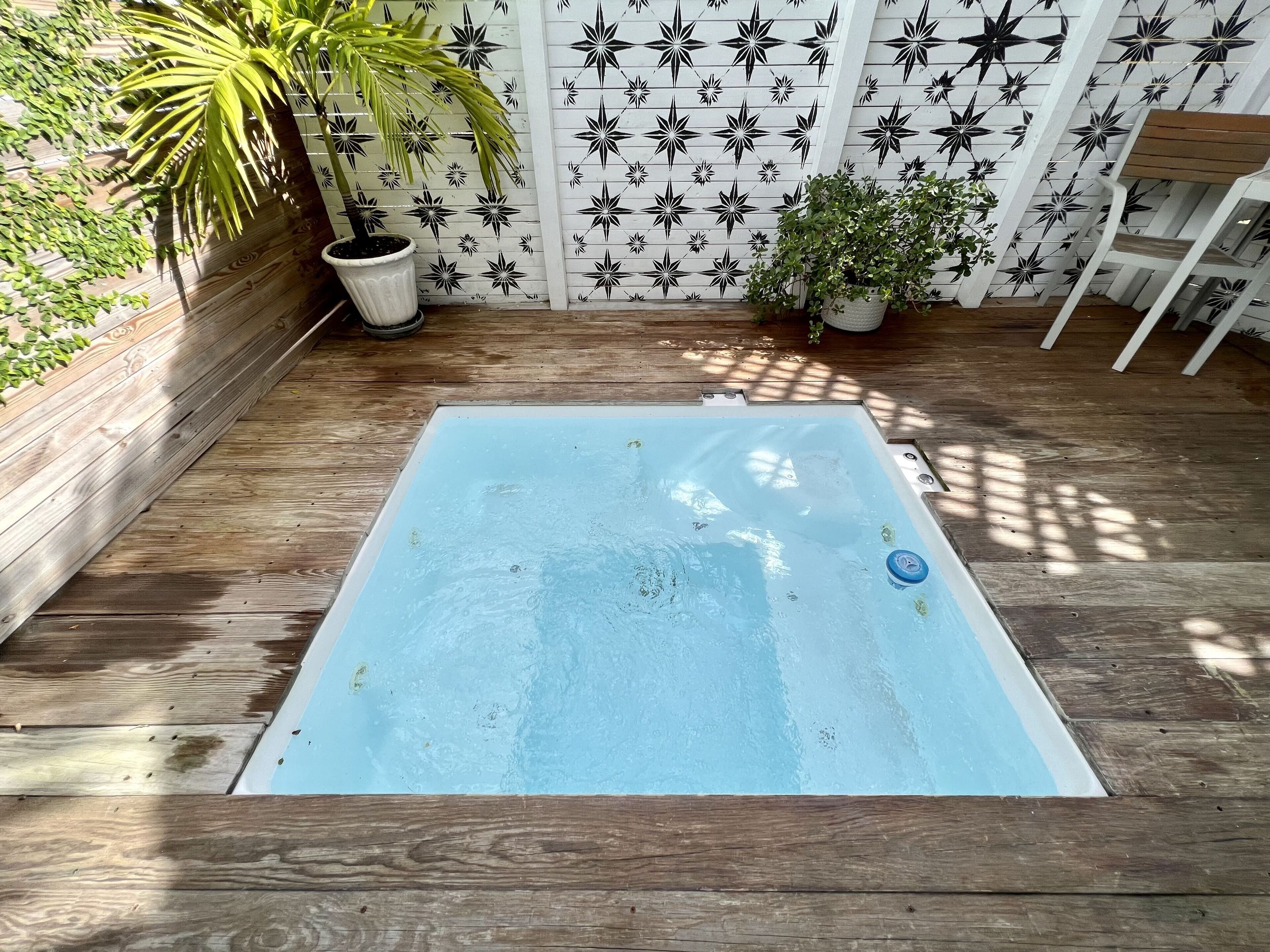 Un bain à remous rectangulaire, avec un intérieur bleu, entouré d’une terrasse en bois et délimité par une clôture blanche ornée de motifs en forme d’étoiles noires.