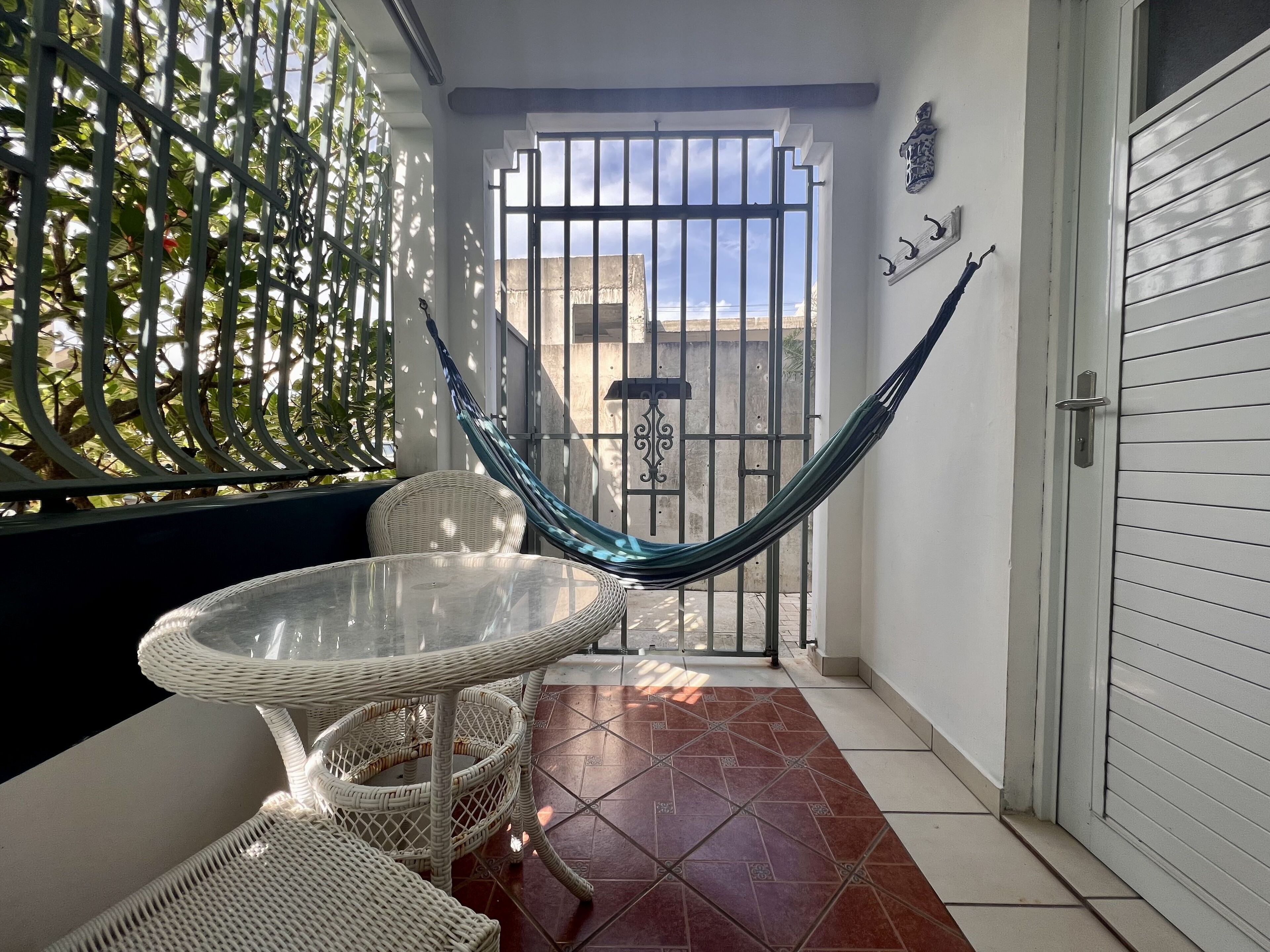 A balcony with a hammock, a glass table, wicker chairs, and a view through a gate.