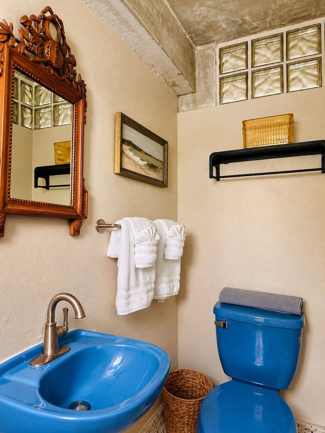 Um banheiro com pia azul, vaso sanitário, espelho com moldura ornamentada, quadro pendurado na parede e porta-toalhas com toalhas brancas.