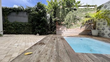 A wooden deck leading to a swimming pool, surrounded by lush greenery and a tiled wall with a star pattern.
