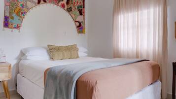 Uma cama bem arrumada com um cobertor bege e rosa, uma mesa de cabeceira de madeira com um abajur, e uma tapeçaria colorida na parede.