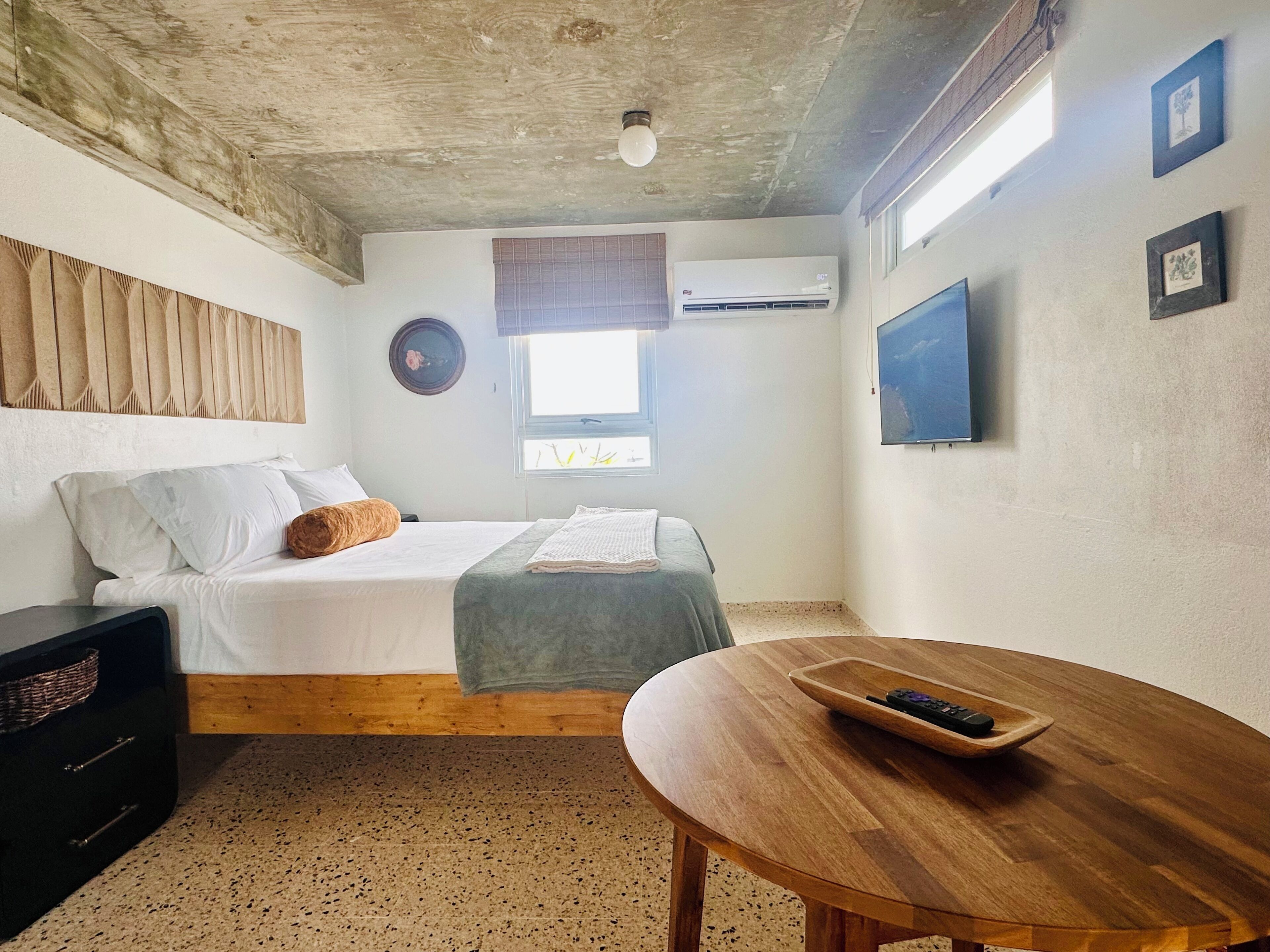 Un dormitorio con una cama, una mesita de noche de madera, una mesa con control remoto, un televisor montado en la pared y un equipo de aire acondicionado.