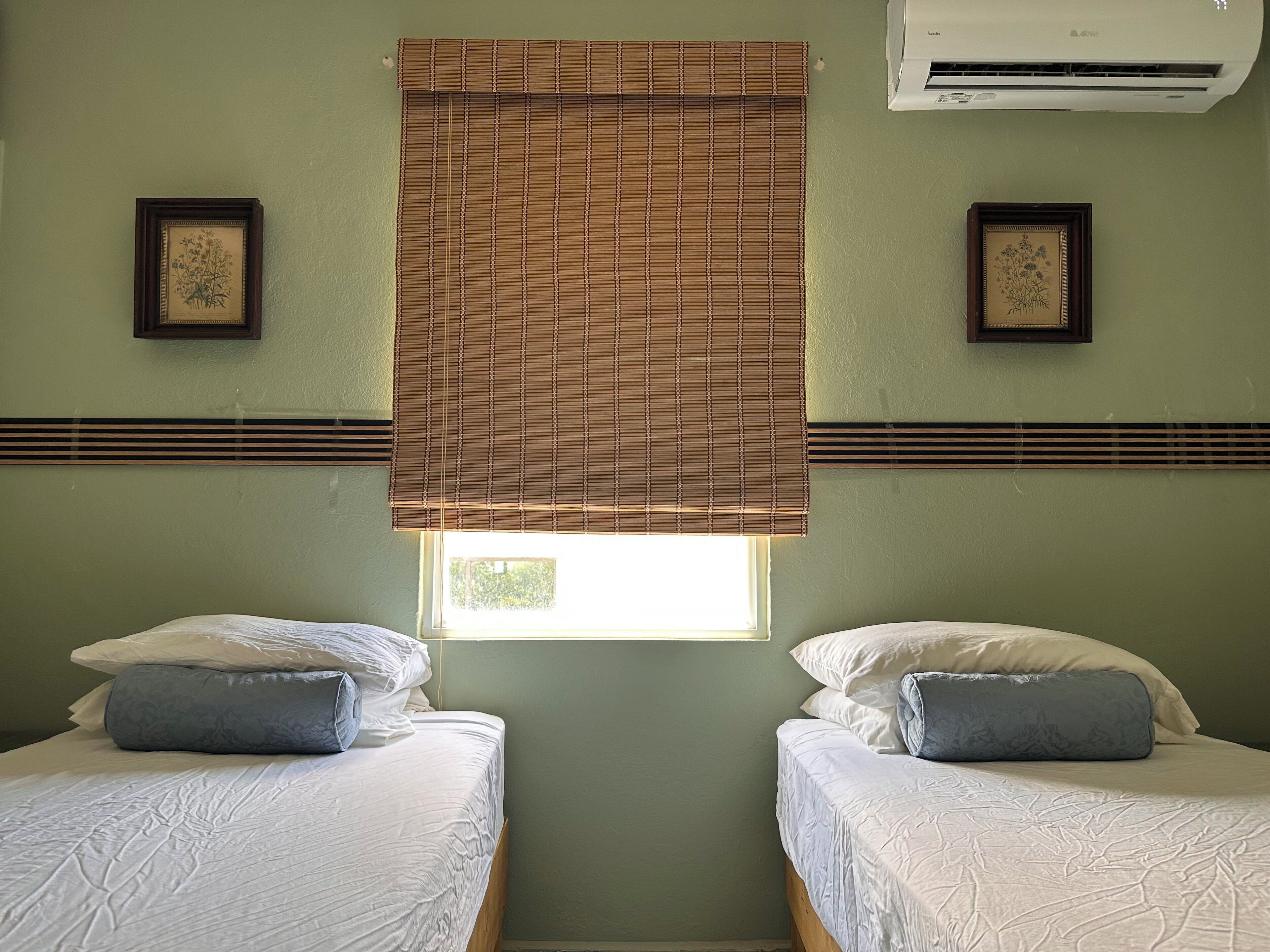 A room with two beds, a window with blinds, an air conditioner, and framed artwork on the walls.