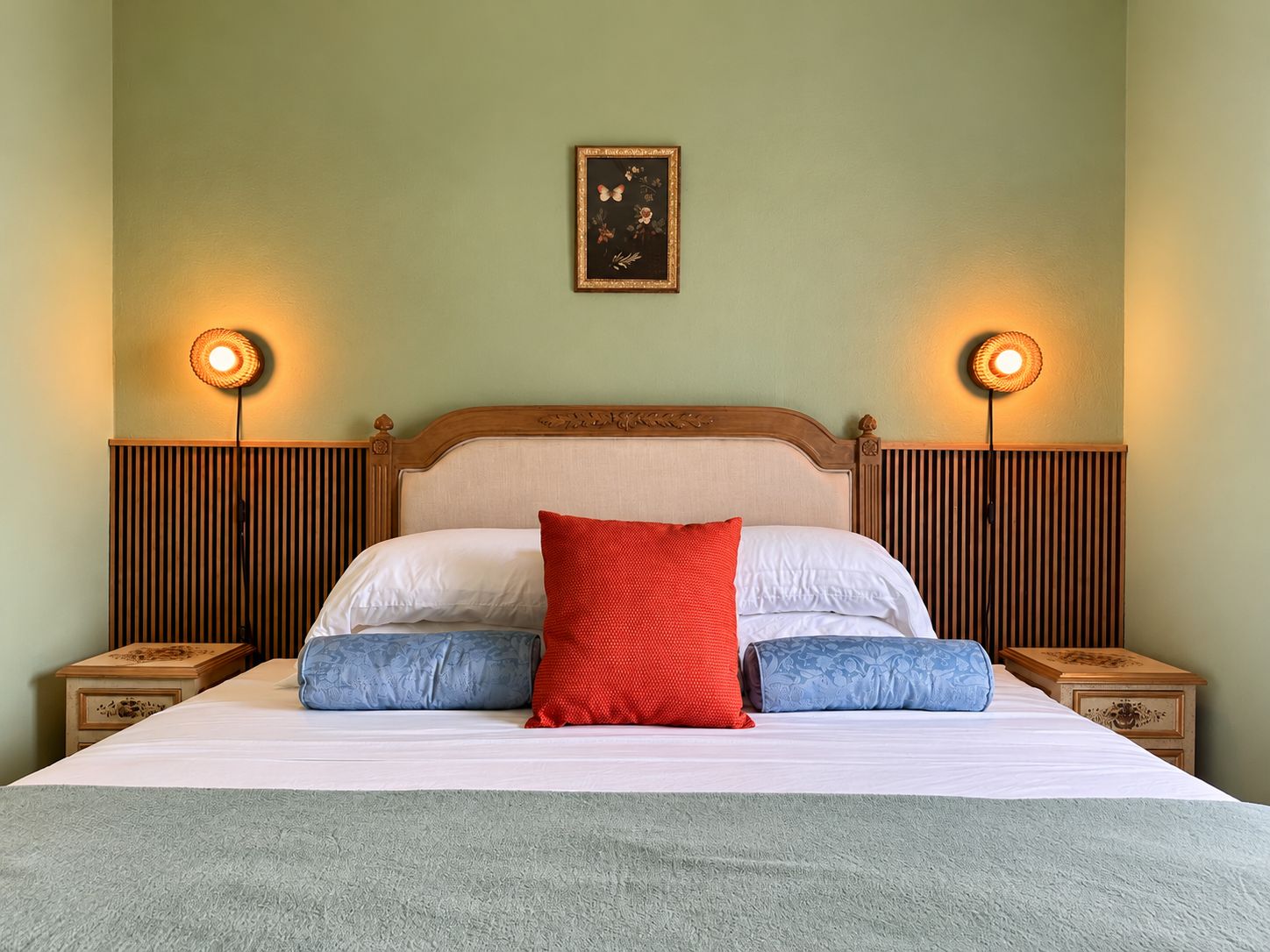Una cama bien tendida con un almohadón rojo, almohadas blancas y almohadones azules. Hay dos mesitas de noche con lámparas a cada lado de la cama.
