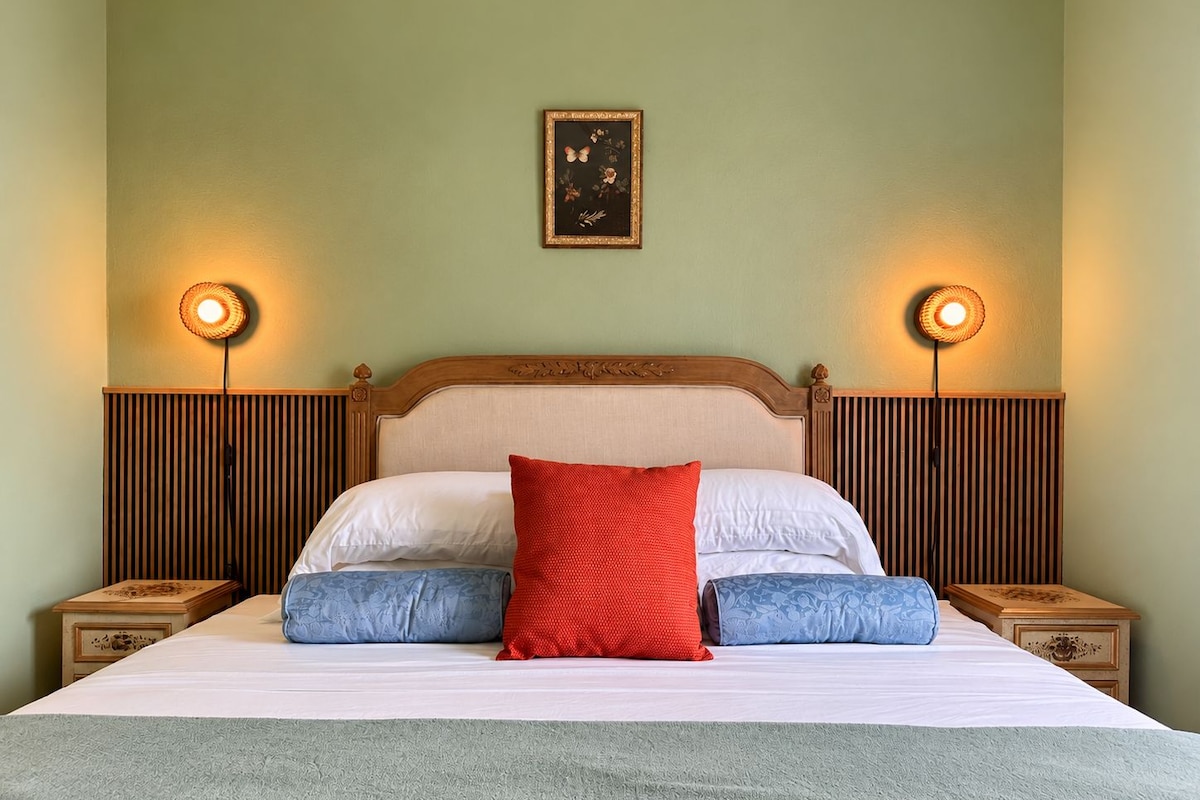 A neatly made bed with a red pillow, white pillows, and blue accent pillows. There are two bedside tables with lamps on either side of the bed.
