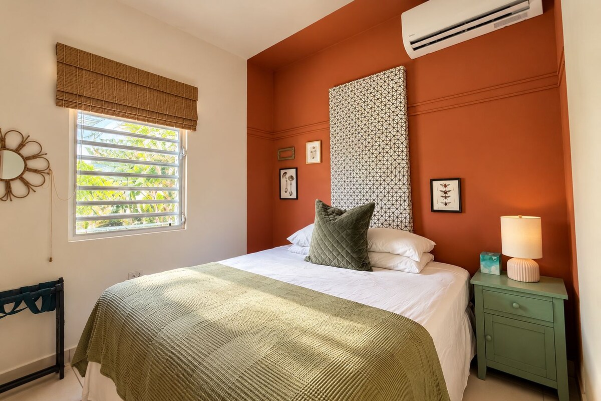 A bedroom with a bed, a bedside table with a lamp, a window with blinds, and an air conditioning unit.