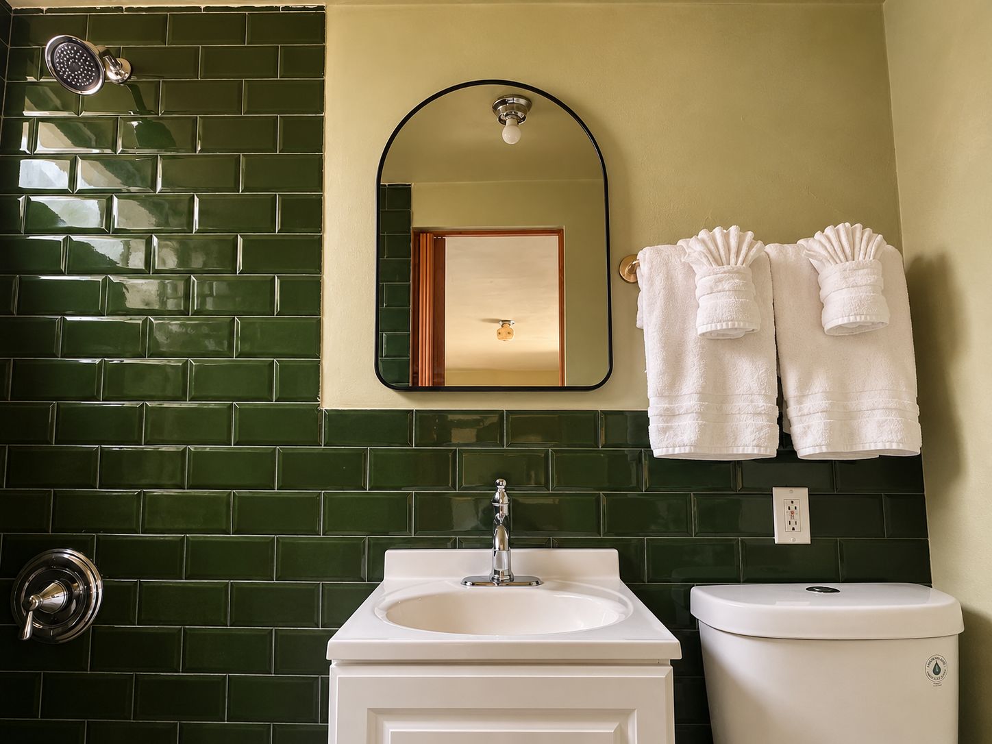 Een badkamer met een groen betegelde muur, een witte wastafel, een toilet, een douchekop, en twee handdoeken die aan de muur hangen.