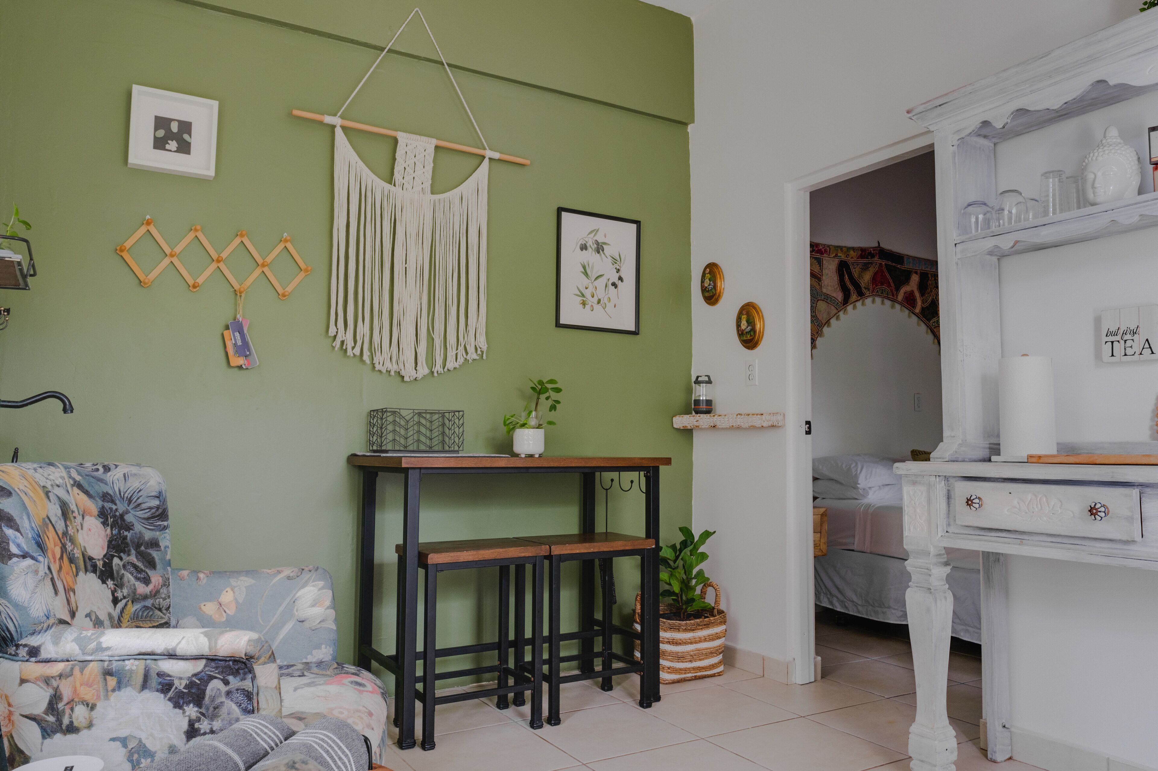 A room with a green wall, a floral sofa, a wooden bench, a white table with a plant, and a white cabinet with tea items.