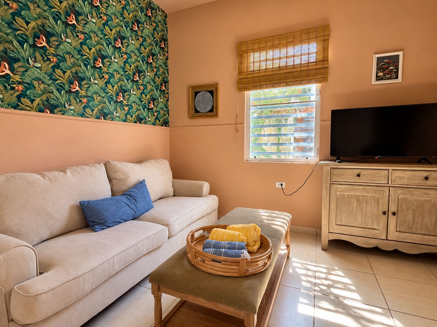 A living room with a sofa, a television, a window with blinds, a wall with a leafy pattern, and a cabinet.