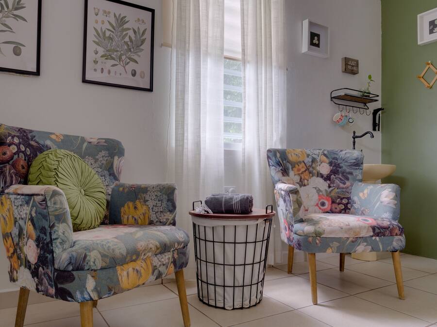 A room with two floral-patterned armchairs, a small round table, and a window with sheer curtains.