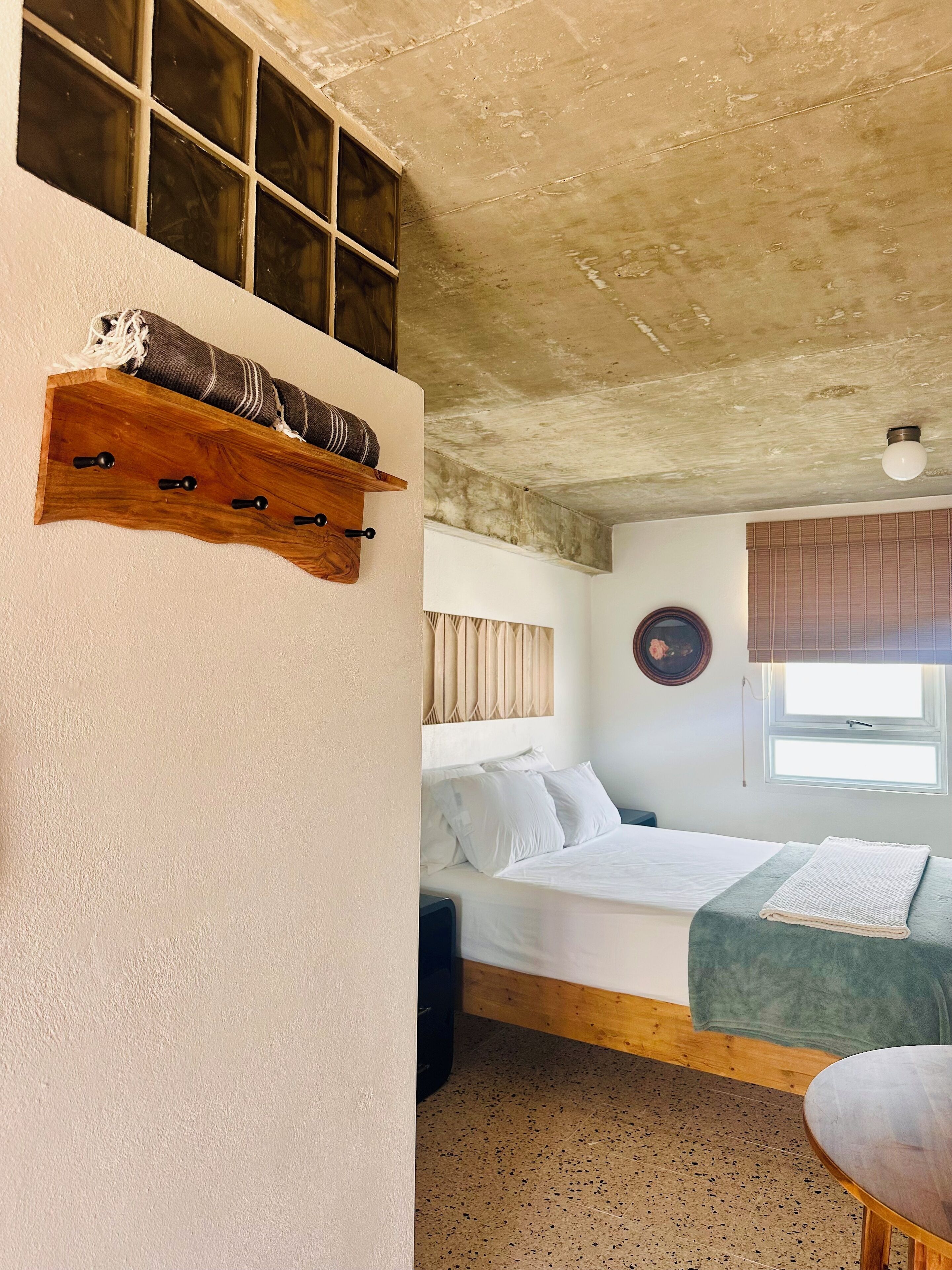 A room with a bed, a bench, a wooden shelf with a blanket, and a window with blinds.