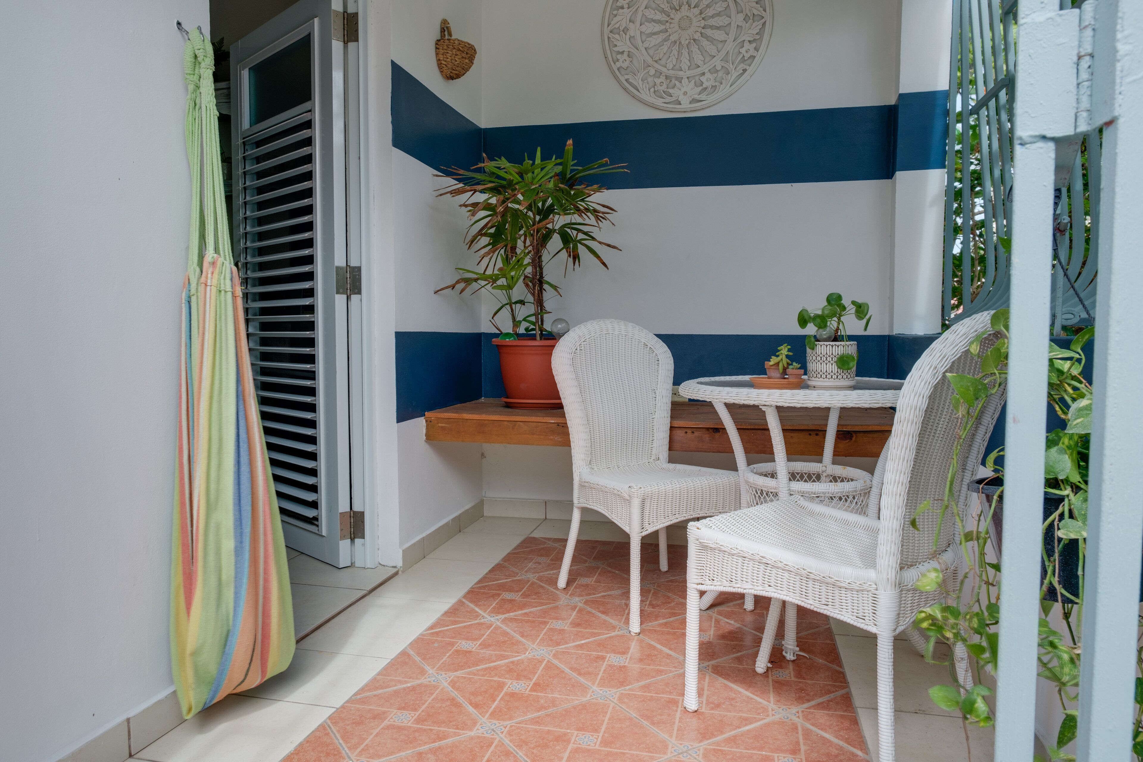 A small patio area with a white wicker table and chairs, a potted plant, and a striped curtain.