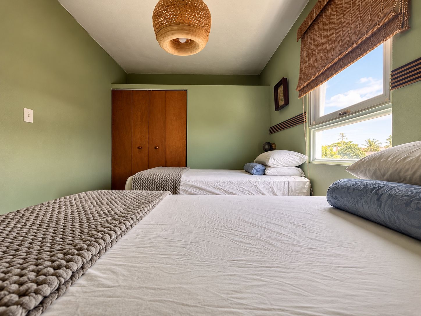 A bedroom with two beds, a wooden wardrobe, a woven ceiling light, and a window with a view.