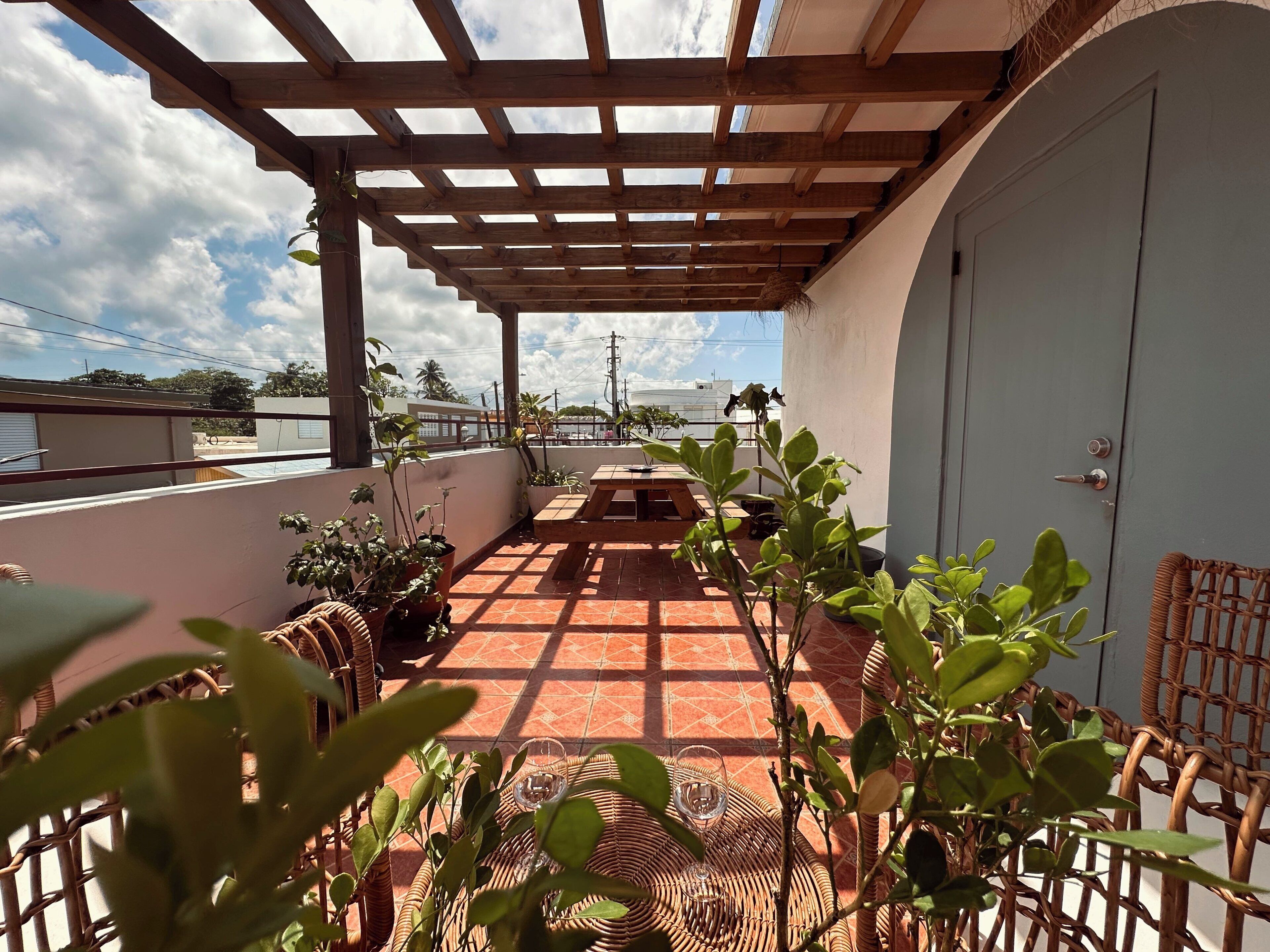 Un patio esterno coperto con una pergola in legno, pavimento piastrellato, mobili in vimini e piante in vaso.