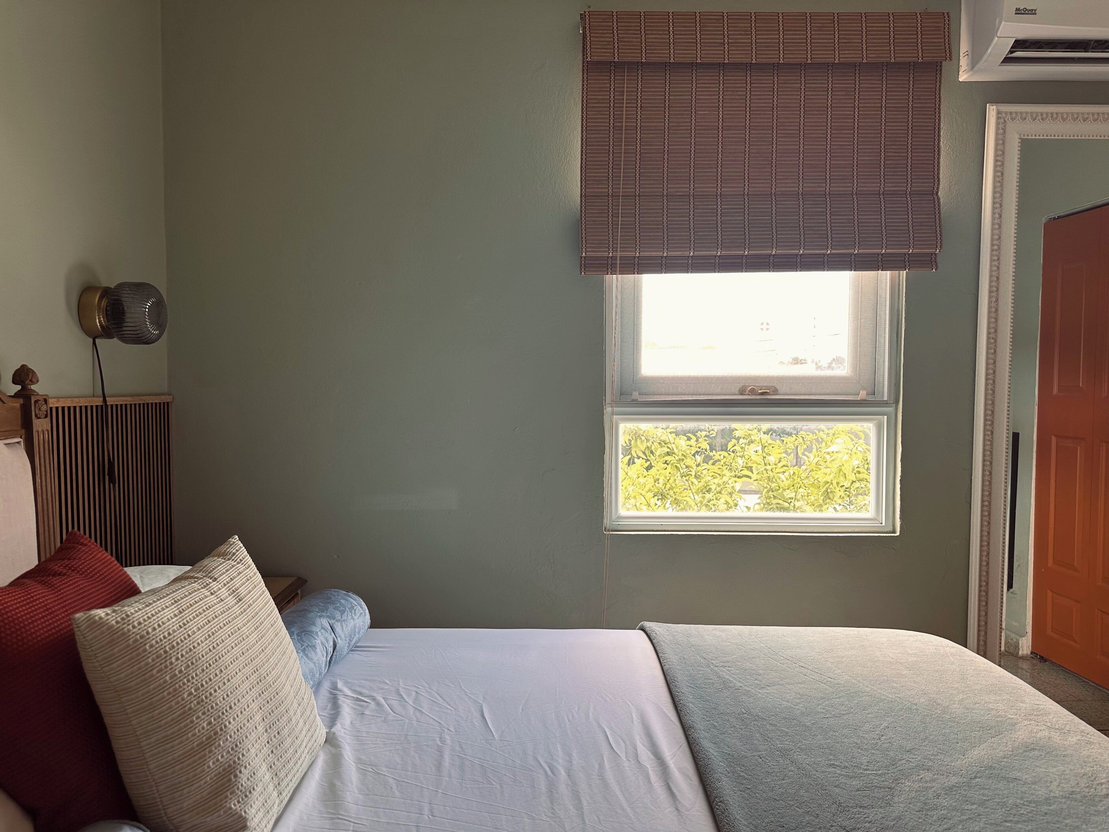 A bedroom with a bed, pillows, a radiator, a window with blinds, and an air conditioning unit.