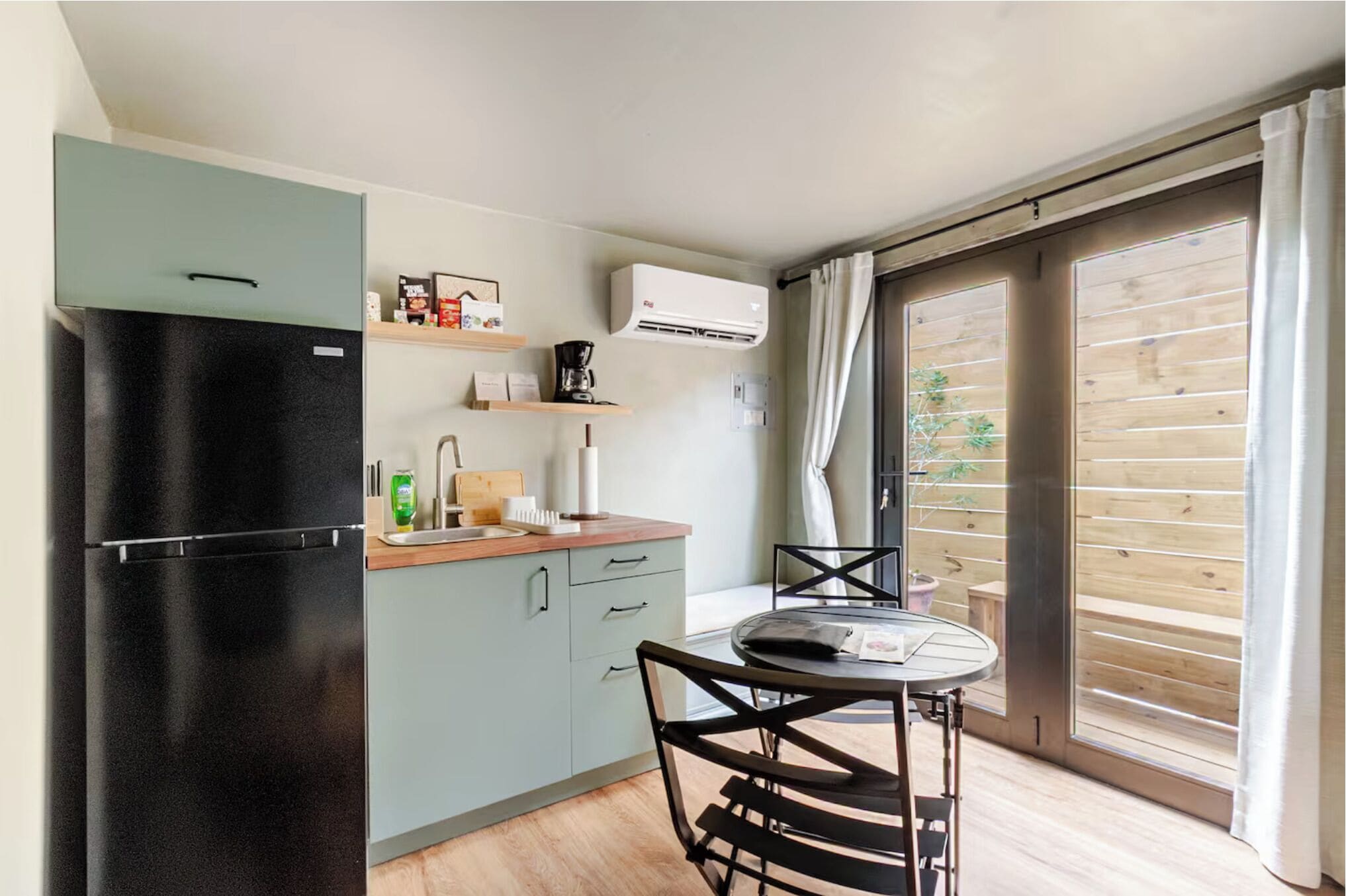 A compact kitchen with a black refrigerator, a wooden countertop, and a dining table with chairs. There is a window with wooden blinds and a view of greenery outside.