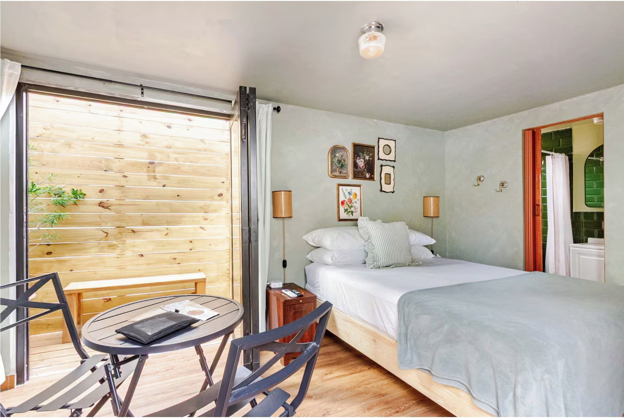 Quarto com cama, mesa de cabeceira, luminária e vista para o exterior através de uma janela com persianas.