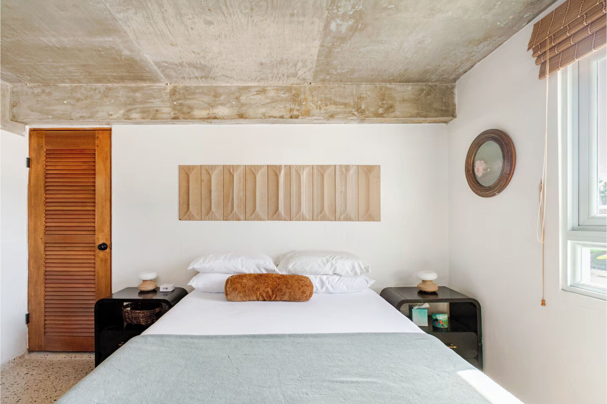 Un dormitorio con cama, cabecera de madera, mesitas de noche, un espejo y una ventana.