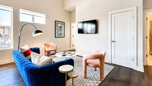 Un salon moderne avec un canapé bleu, un fauteuil rose, une table basse ronde et un téléviseur à écran plat fixé au mur.