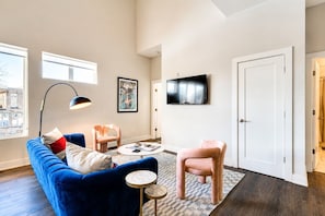 Una sala de estar moderna con un sofá azul, un sillón rosa, una mesa de centro redonda y un televisor de pantalla plana montado en la pared.