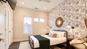 Une chambre à coucher avec un lit, deux lampes de chevet, un miroir rond, un fauteuil en osier et une fenêtre donnant sur des immeubles.