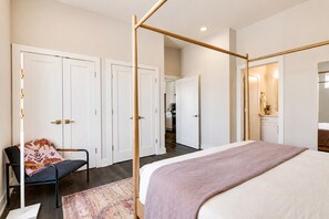 Een slaapkamer met een hemelbed, een stoel met een gekleurd kussen, en een gang naar een badkamer.