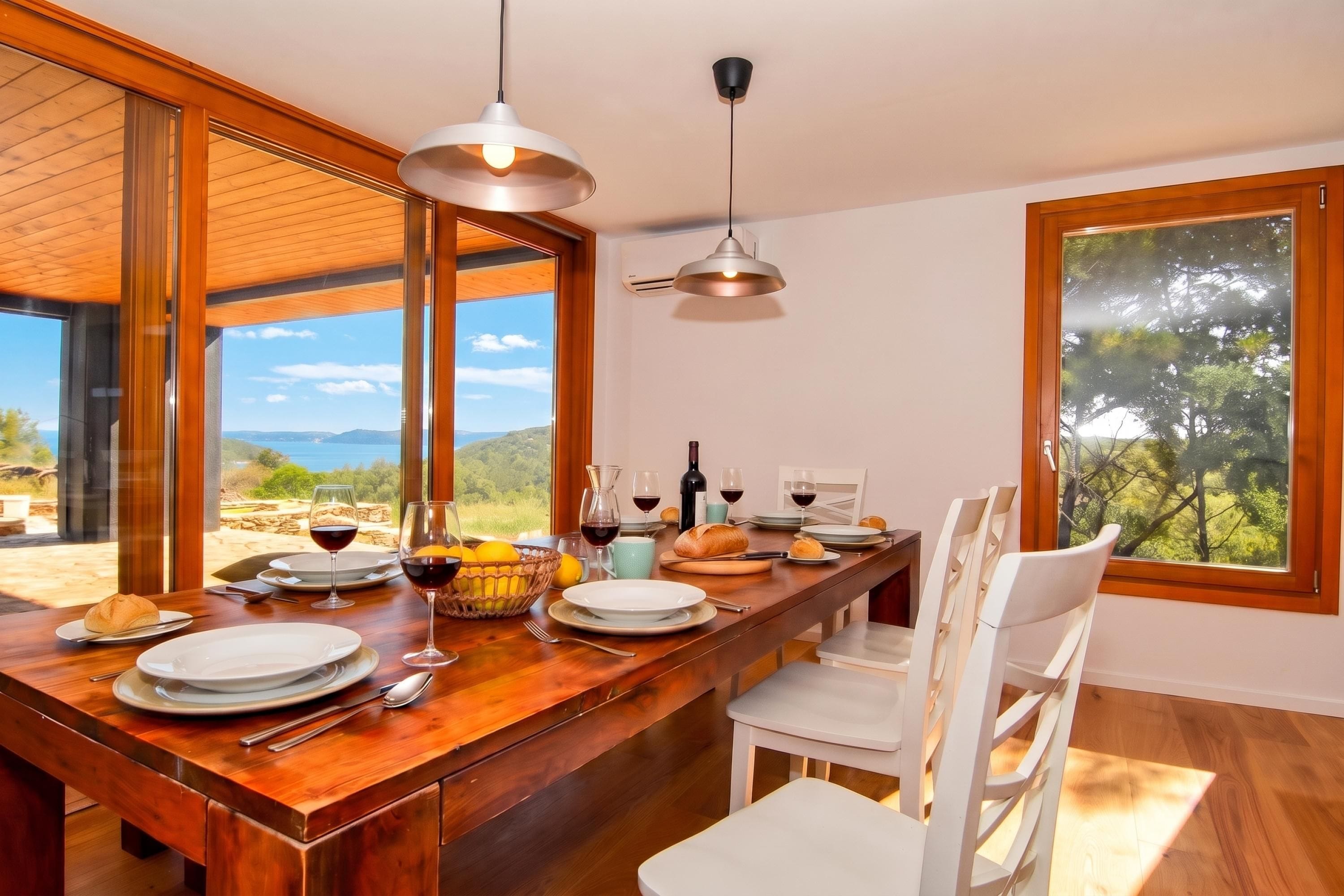 Uma área de refeições com mesa de madeira posta para uma refeição, grandes janelas com vista para o exterior e um moderno lustre.