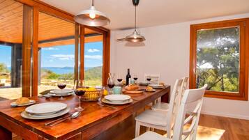 Uma área de refeições com mesa de madeira posta para uma refeição, grandes janelas com vista para o exterior e um moderno lustre.