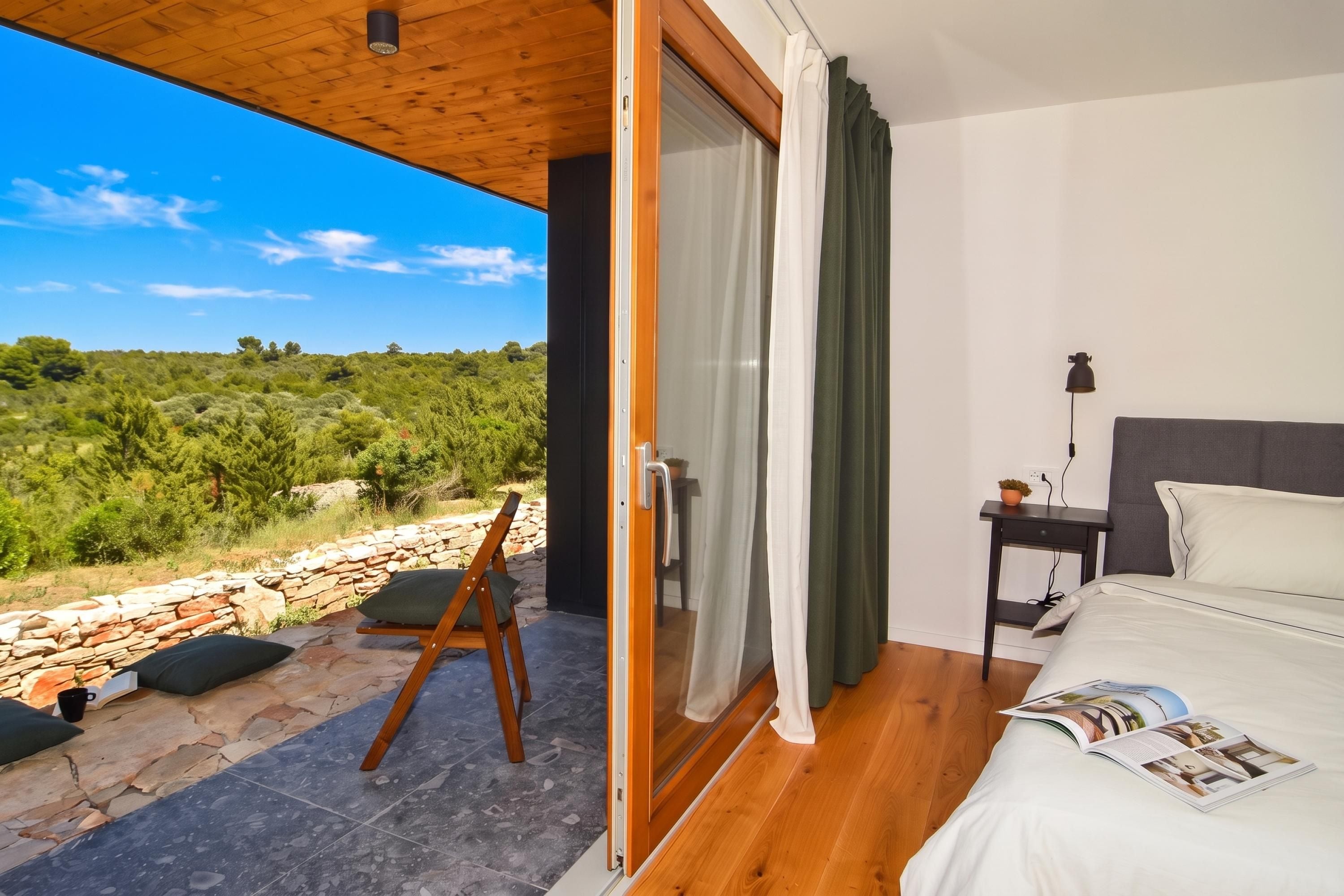 A bedroom with a bed, a nightstand, a lamp, and a view of the outdoors.
