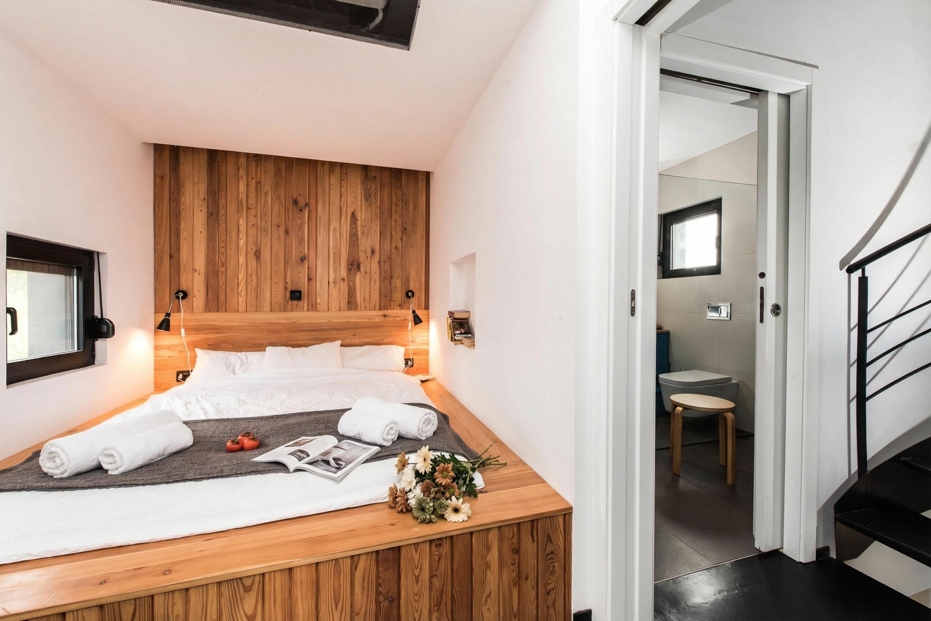 A bedroom with a wooden headboard, a bed with white linens, a bedside table with a magazine and flowers, and a view of a bathroom through an open door.