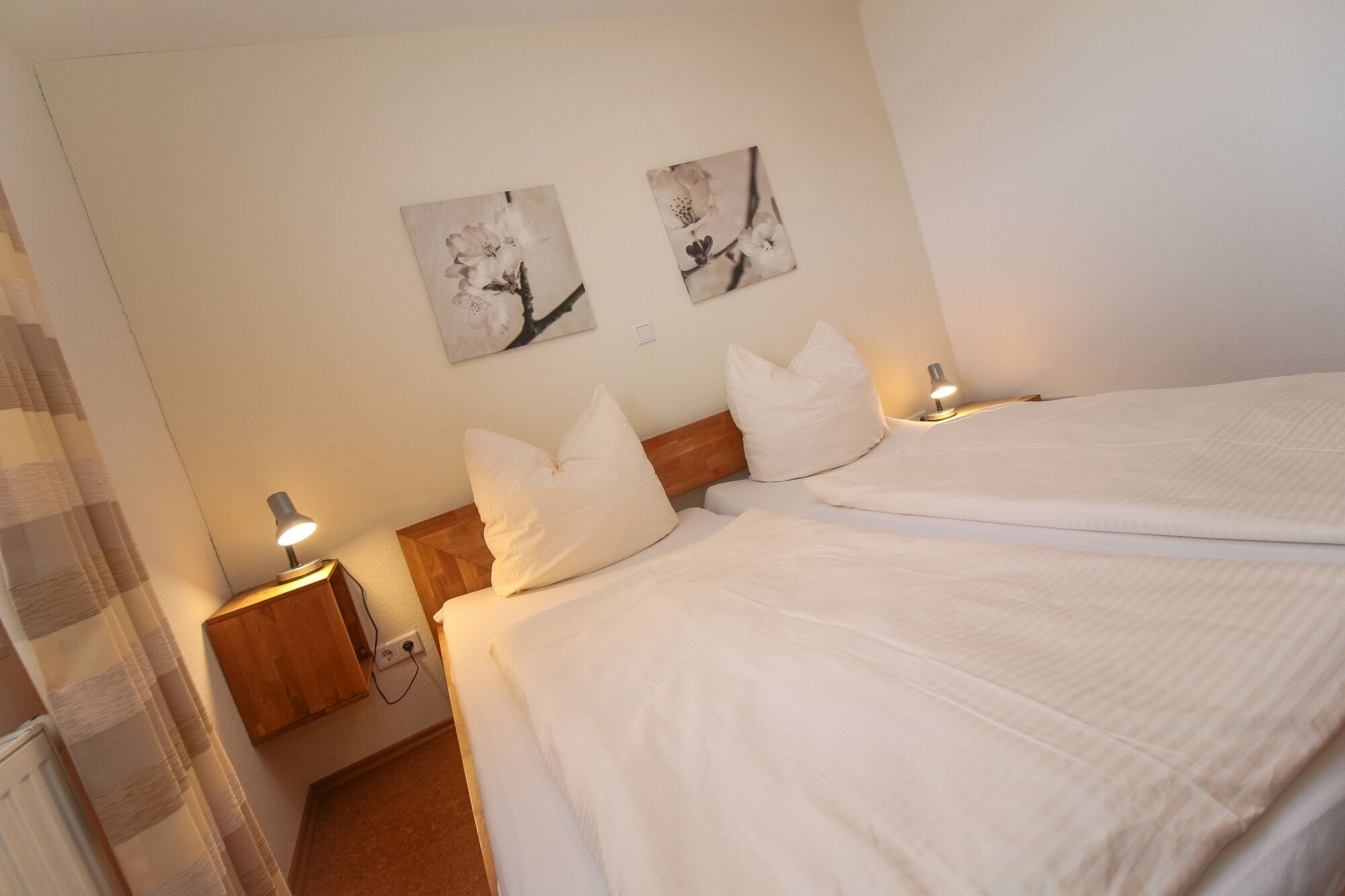 Quarto de hotel com duas camas, roupa de cama branca, cabeceira de madeira, abajures e dois quadros florais fixados na parede.