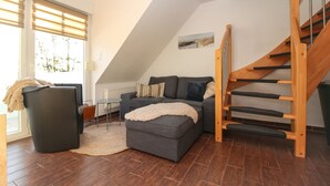 A modern living room with a grey sofa, a small ottoman, a woven rug, and a wooden staircase. - Ferienwohnung Oberdeck (Norden)