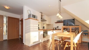 A modern kitchen with white cabinets, a microwave, and an oven. There is a dining area with a wooden table and white chairs. A TV is mounted on the wall above the dining area. - Ferienwohnung Oberdeck (Norden)