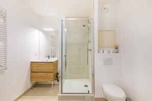 Un baño moderno con ducha envuelta en vidrio, tocador de madera, inodoro y un estante pequeño con artículos de aseo.