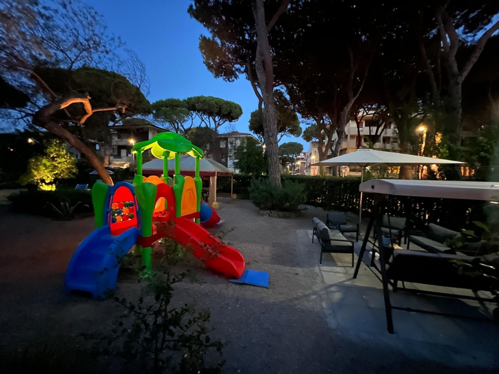 Ein Spielplatz mit Rutsche und Klettergeräten, umgeben von Bäumen und Sitzgelegenheiten im Freien.