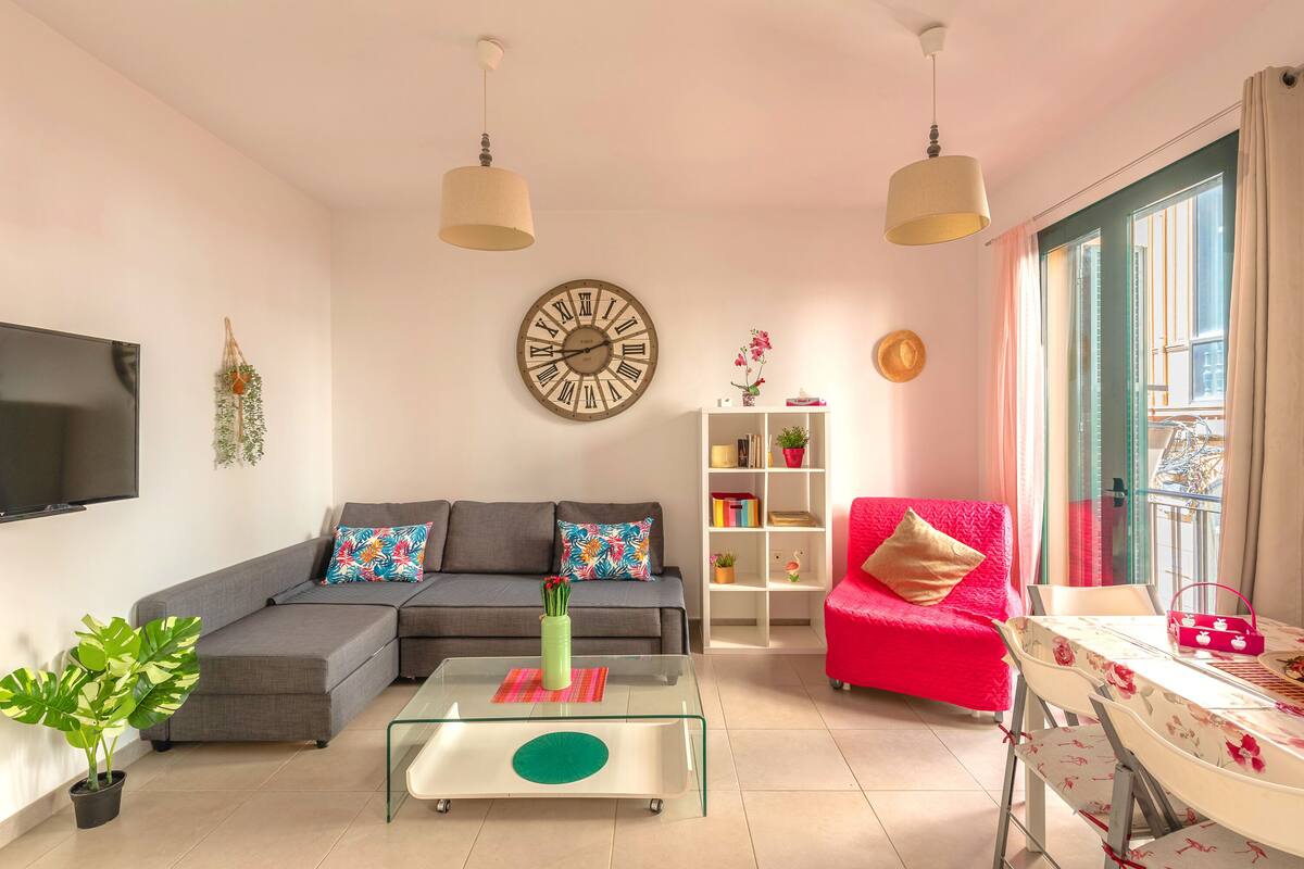 Uma sala de estar com sofá seccional, mesa de centro de vidro, poltrona vermelha, estante branca e um relógio de parede grande.