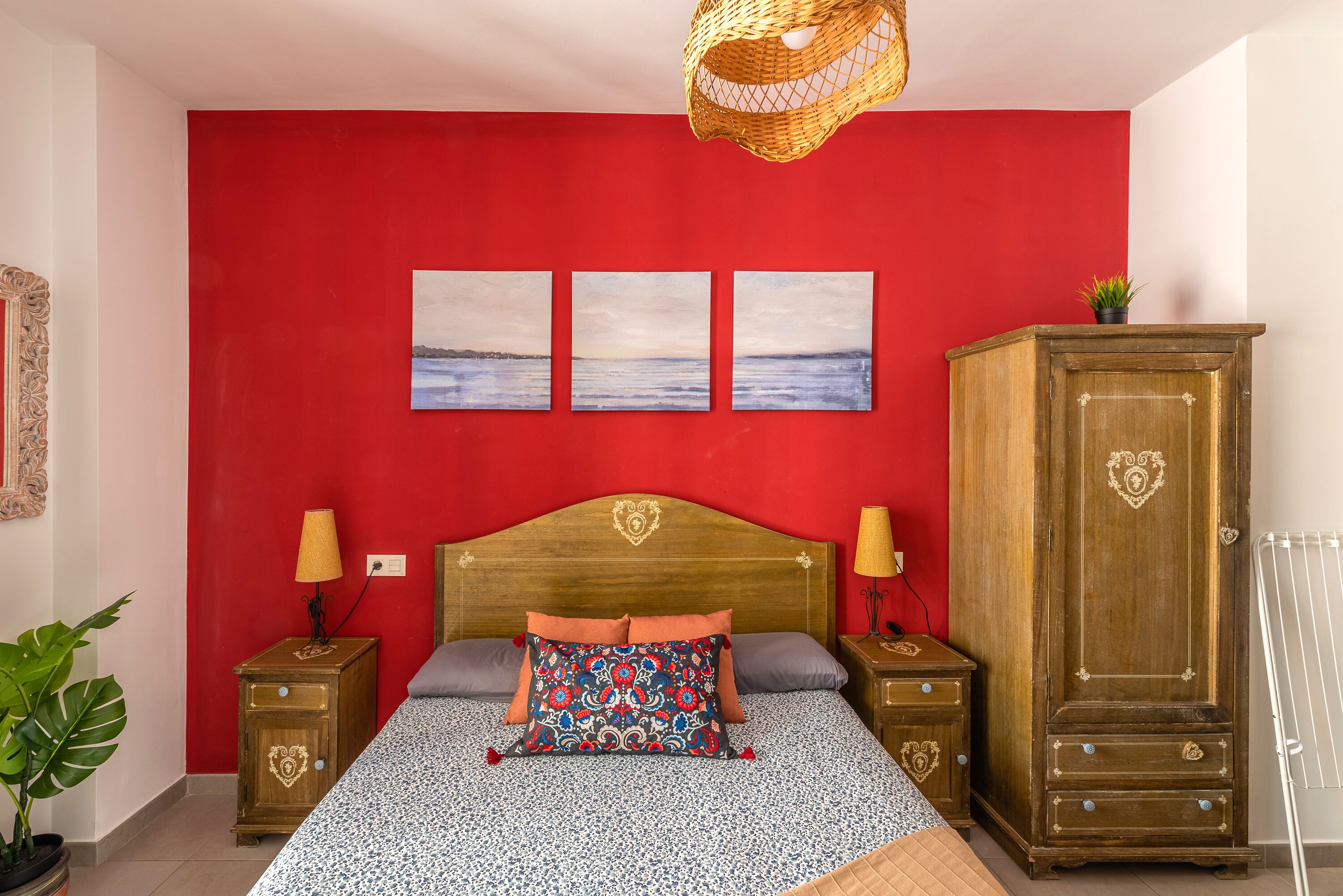 Een slaapkamer met een rode wand, een houten hoofdbord, twee nachtkastjes met lampen, een groot bed met beddengoed met een patroon, en een garderobe.