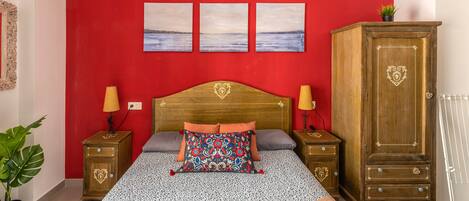 Een slaapkamer met een rode wand, een houten hoofdbord, twee nachtkastjes met lampen, een groot bed met beddengoed met een patroon, en een garderobe.