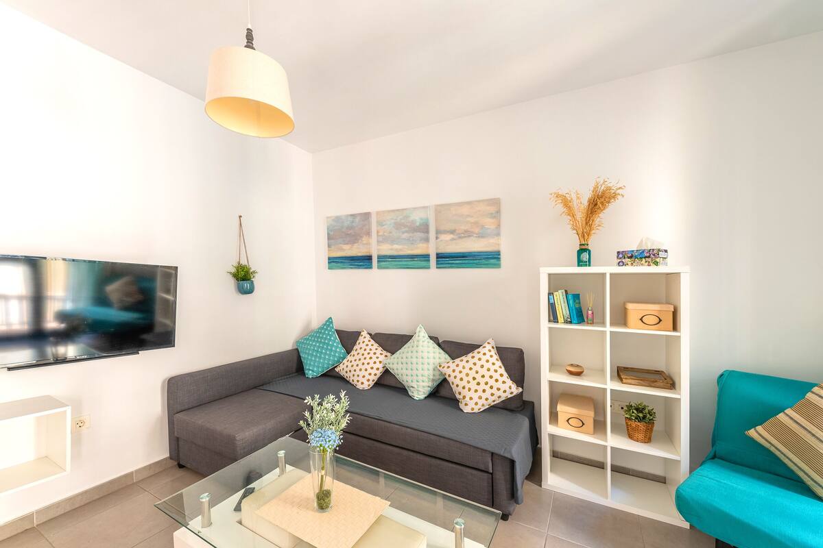 Uma sala de estar moderna com um sofá cinzento, uma poltrona azul-turquesa e uma mesa de centro de vidro. Uma televisão de tela plana está fixada na parede, e há almofadas decorativas e um móvel de estantes branco com caixas organizadoras.