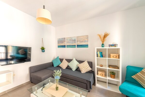 A modern living room with a grey sofa, teal armchair, and a glass coffee table. A flat-screen TV is mounted on the wall, and there are decorative pillows and a white shelving unit with storage boxes. - Skysea Holidays City Center Suites (Málaga)
