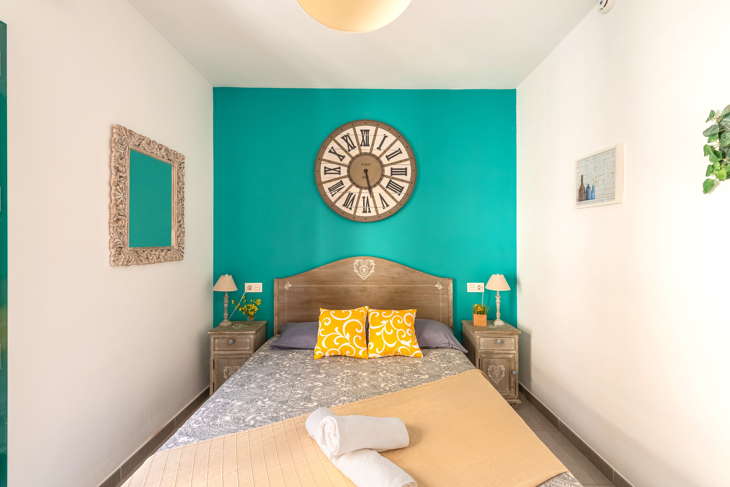 Ein Schlafzimmer mit einem Bett, einer großen Wanduhr, einem Spiegel und einem kleinen Tisch mit einer Lampe.