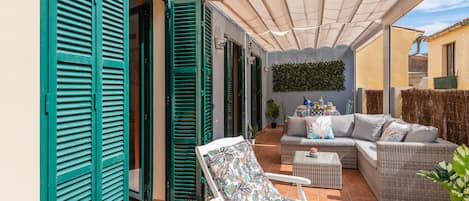 A covered patio with wicker furniture, a white chair with a floral cushion, and a sofa with cushions.